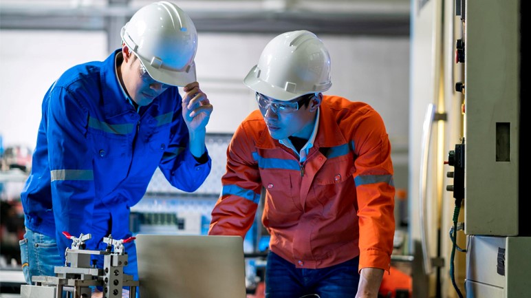 Two men in hard hats collaborate on a laptop, focused on their work in a construction or industrial setting.